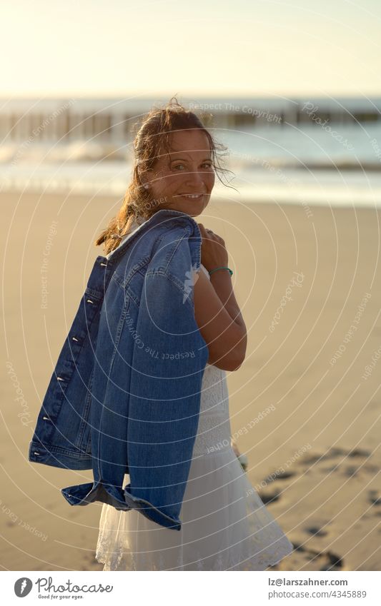 Attractive middle-aged woman in stylish white dress carrying a bouquet of flowers and jacket slung over her shoulder walking across a deserted beach at sunset turning to smile at the camera