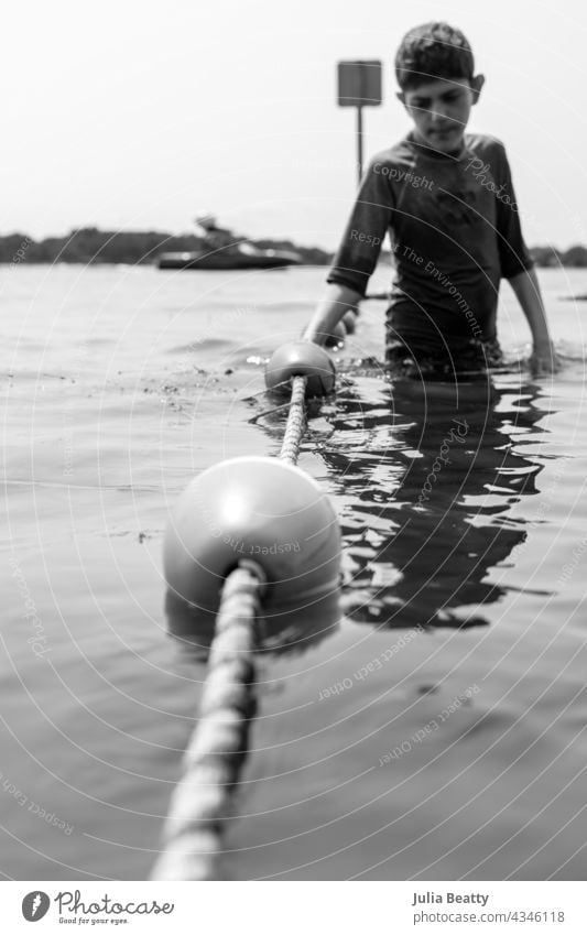 Young boy holding onto rope barrier at lake; buoys on rope wade safety pre-teen autism special needs isolated solitary beach summer water nature vacation