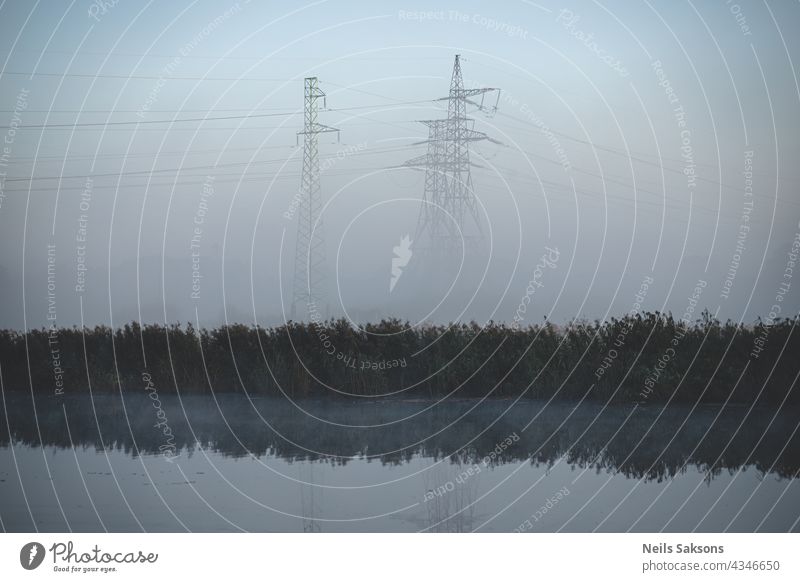 electric transit lines and pylons near river in misty morning in Latvia. Lots of dry reeds in october reflected in water. Autumn cold morning fog in background.