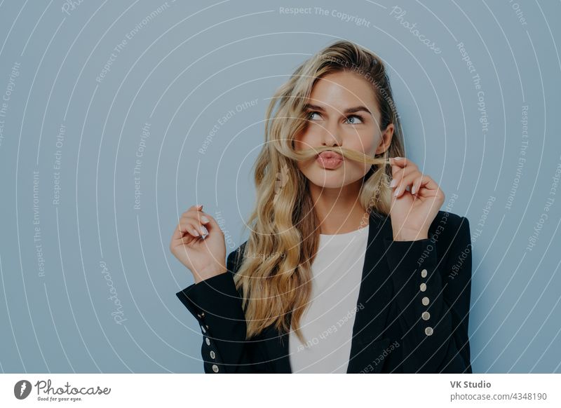 Funny joyful blonde woman having fun while posing against blue studio wall, playing with strand of hair funny carefree happy beautiful young white pretty