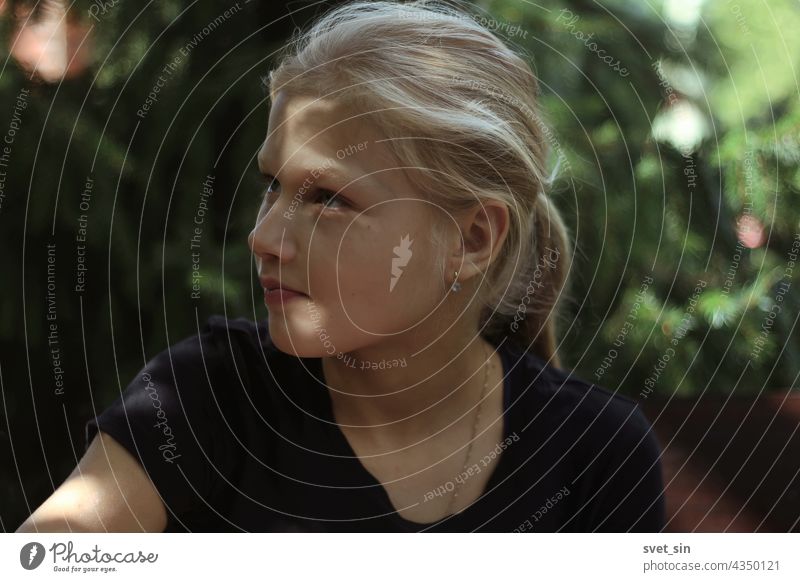 A blond teenage girl in a black T-shirt is sitting outdoors in the greenery and looking away with interest. Portrait of a pretty blonde girl in a semi-profile with sun glare on her face.