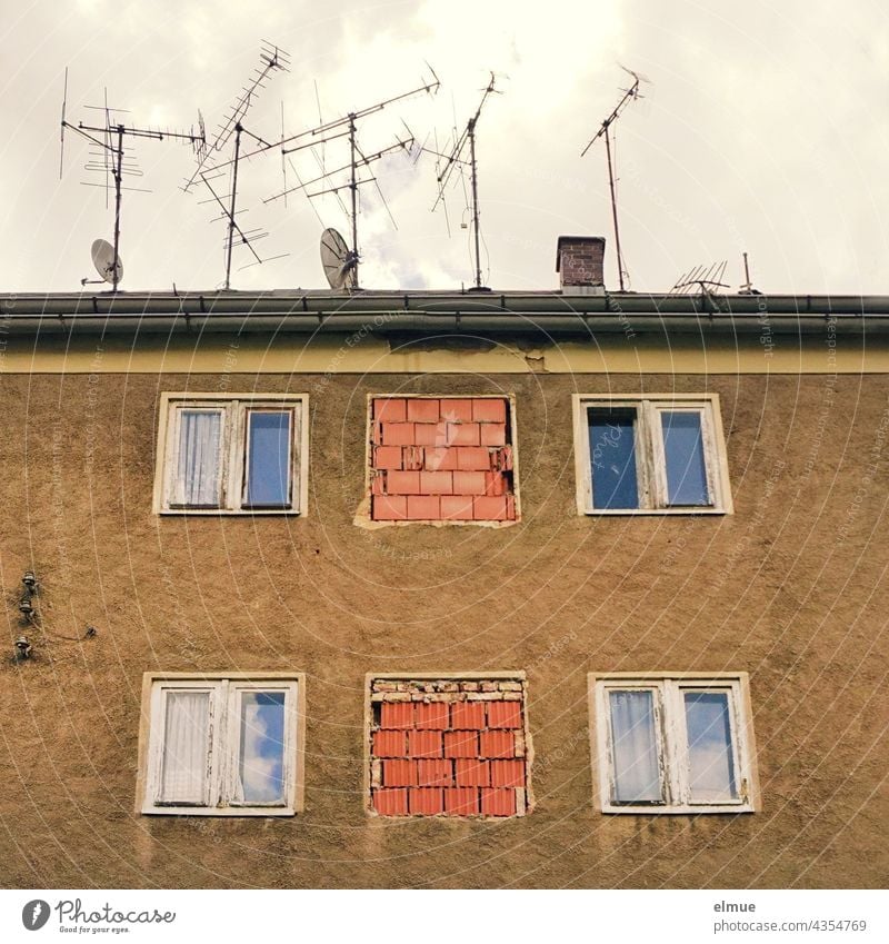 View of an old, unoccupied apartment building with windows, partly bricked up, and numerous old TV antennas as well as satellite dishes on the roof / dilapidated / core renovation / real estate