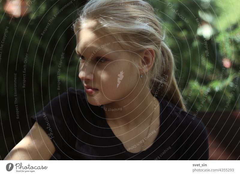 A blond teenage girl in a black T-shirt is sitting outdoors in the greenery and looking thoughtfully aside. Portrait of a pretty blonde girl in a semi-profile with sun glare on her face.