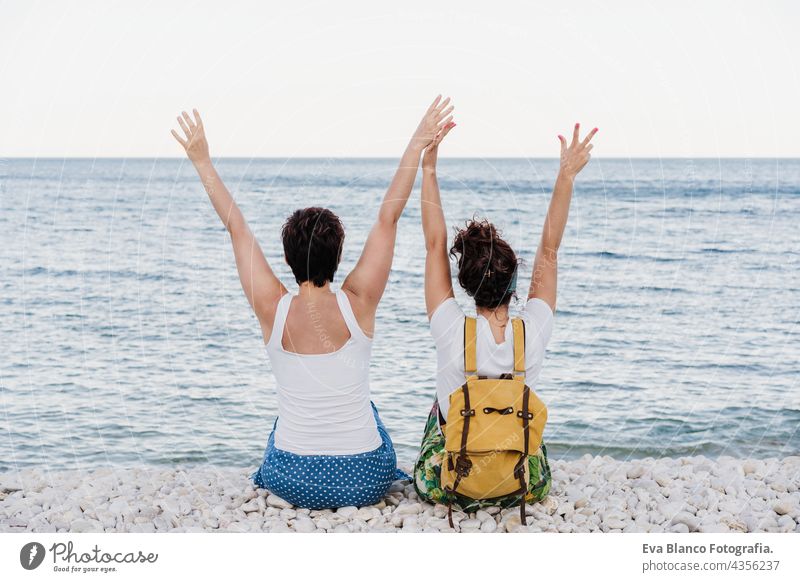 back view of happy lesbian couple sitting at the beach with arms raised during sunset. Love is love and LGTBI concept women homosexual summer together