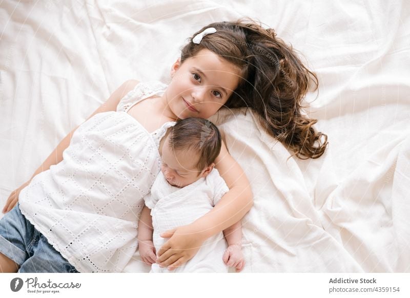 Smiling girl lying with newborn on bed child together sibling baby infant hug kid cute adorable childhood home innocent little happy embrace babyhood joy