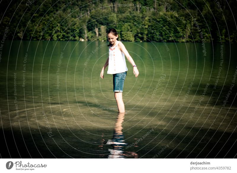 A young girl in the cold Königsee. Very carefully she goes into the green shimmering cold dark lake with her feet. In the background the overgrown steep slopes