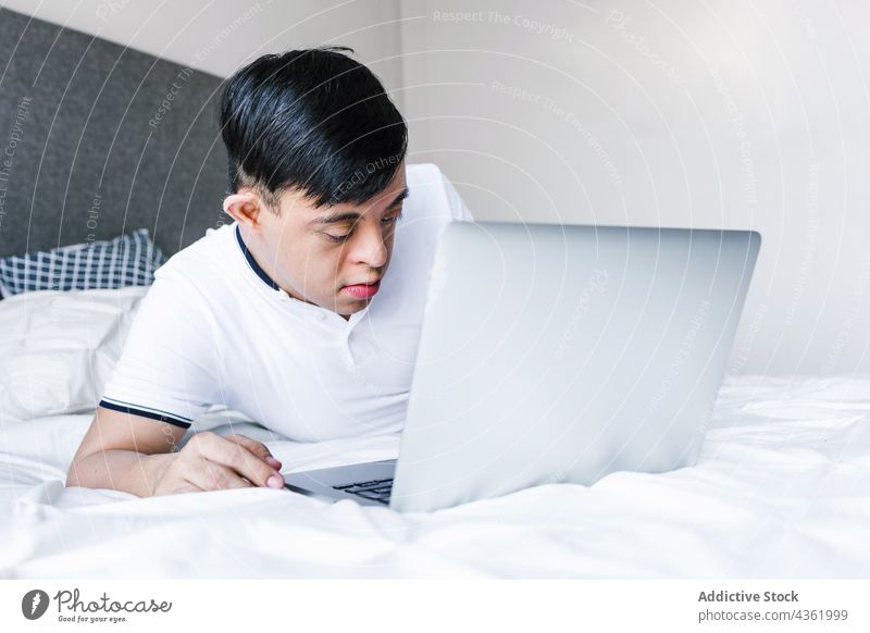 Ethnic teenage boy with Down syndrome browsing laptop at home down syndrome lying bed surfing using ethnic latin entertain gadget internet concentrate device