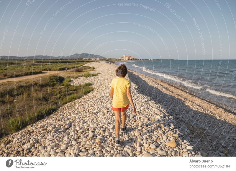 A cheerful and vital woman, in her 70s, walks along a natural pebble beach at sunset, by the Mediterranean Sea, Spain take a walk walking on the beach