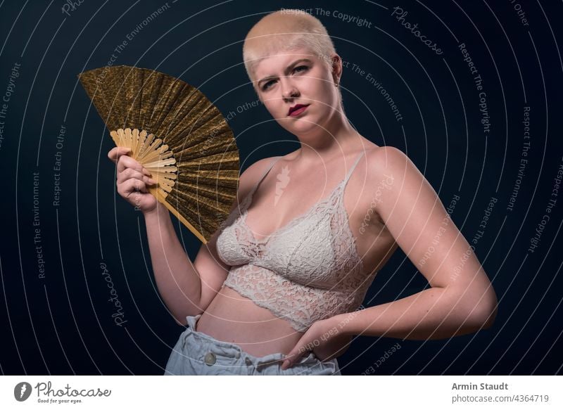 studio shot of a young, strong woman with very short blonde hair, a fan and a bustier serious confident power powerful business piercing jewelry portrait female