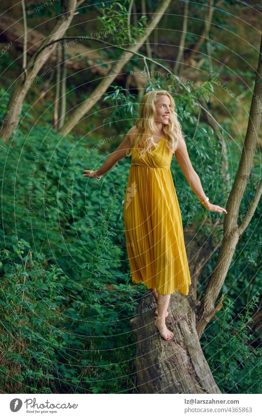 Carefree barefoot attractive blond woman balancing on an old fallen log in a forest wearing an elegant yellow dress looking away to the side with a happy friendly smile