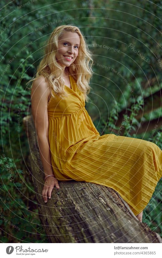 Attractive blond woman in a stylish yellow dress posing on an old tree trunk against greenery in summer looking at the camera with a smile in a close up portrait