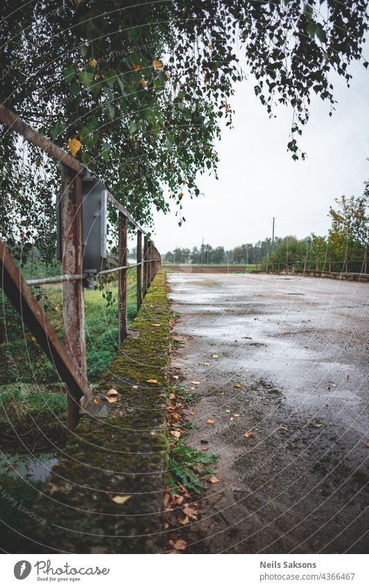 old vintage concrete bridge in Latvia over river Iecava. Iron welded railing. Birch branch with green and yellow trees. Its early september evening, dull grey sky and small rain. Asphalt surface.