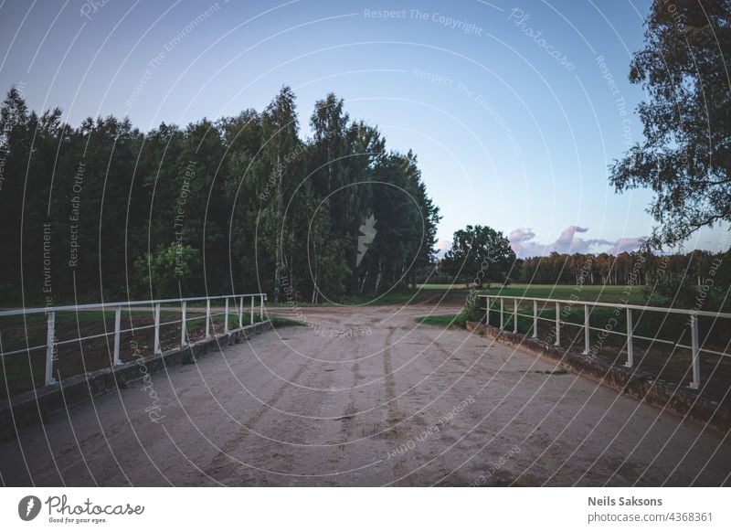 concrete bridge with metal railing over river leads to T type crossroad. Forest and green meadow in distance, blue sky, evening sunset light architecture