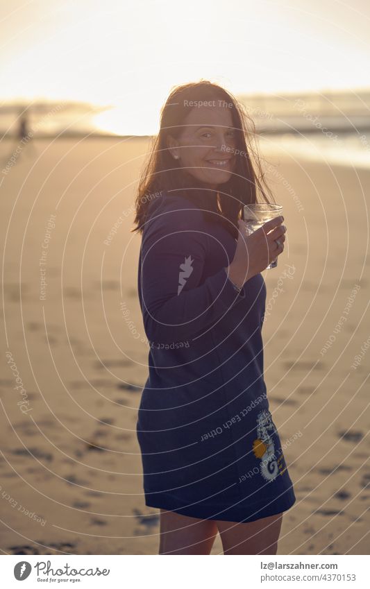 Woman enjoying sunset on a tropical beach while drinking a cocktail turning to look over her shoulder at the camera with a happy vivacious smile woman brunette