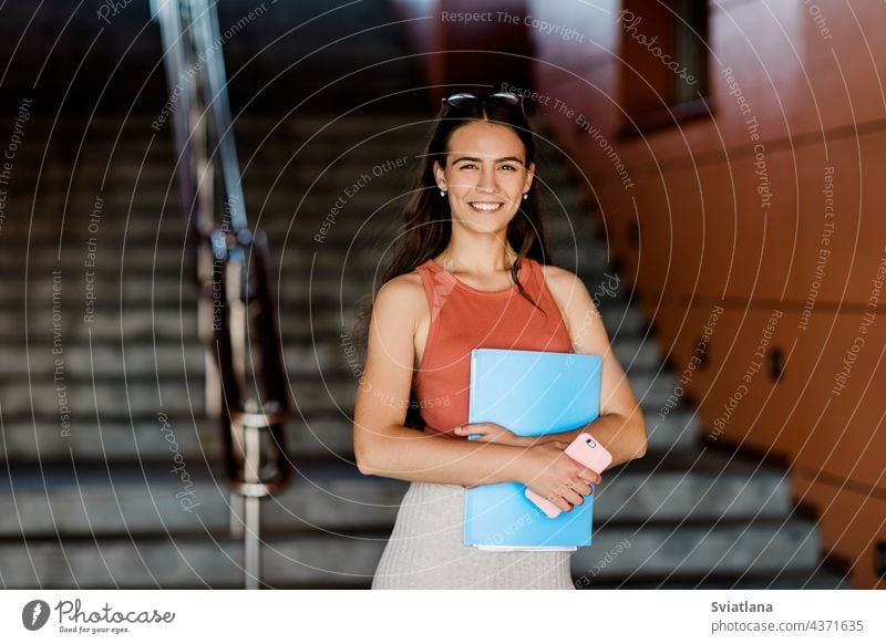 A young student in casual clothes with an essay in a folder and a phone in her hands girl glasses stairs woman smartphone university female adult portrait