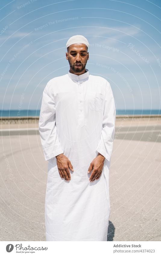 Muslim man praying on beach muslim arab islam religion tradition culture rug ritual worship holy male ethnic authentic sand arid desert blue sky cap robe faith