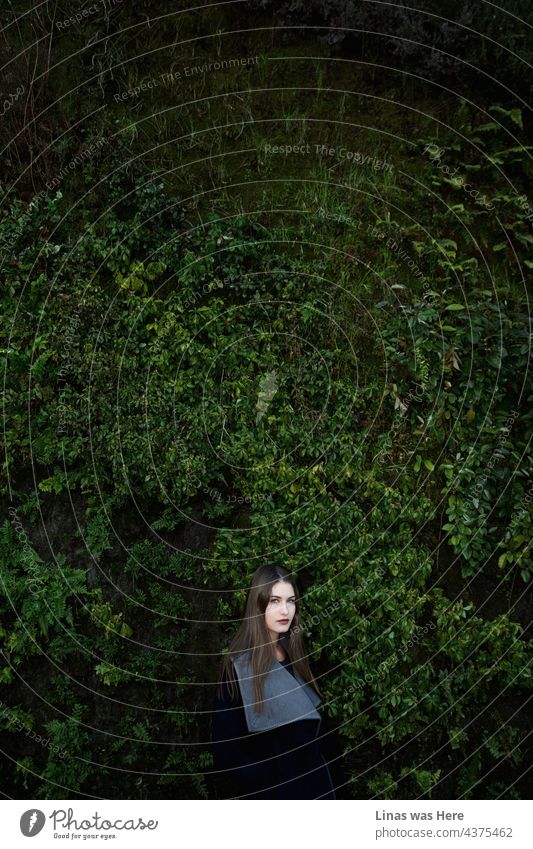 A gorgeous brunette girl is standing right next to a green wall in Madrid, Spain. Wearing a fashionable coat, red lipstick, and looking straight into the camera. Her eyes are piercing through the lens of a camera undoubtedly.