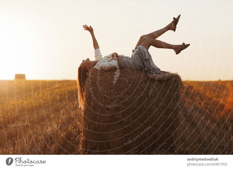 Young woman with long hair, wearing jeans skirt, light shirt is lying on straw bale in field in summer on sunset. Female portrait in natural rural scene. Environmental eco tourism concept.