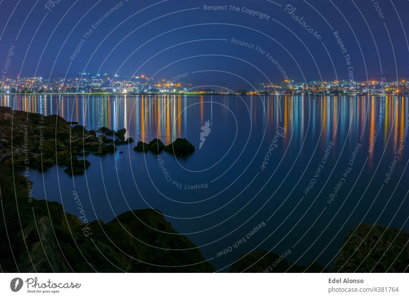 Night view of a bay with dark rocks in the foreground and in the background the illuminated city which colored lights are reflected in a calm sea. The atmosphere is deep blue and quiet.