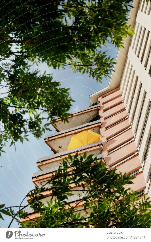 Summer in the city - balconies and facade of a skyscraper from below, a bright yellow parasol and the green leaves of the trees in front of it at home Gray