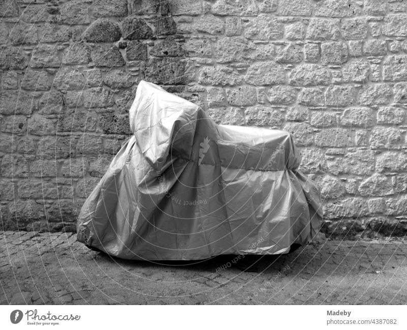 Tightly fitting tarpaulin as if by a packaging artist over a motor scooter in front of old masonry in the alleys of Alacati at the Aegean Sea in the province of Izmir in Turkey, photographed in classic black and white