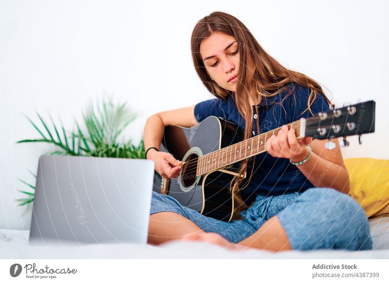 Young woman with laptop playing guitar music learn online at home acoustic practice female student young hobby lesson tutorial instrument watch education using