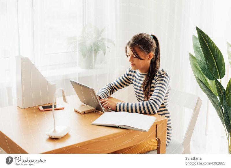 A dark-haired schoolgirl sits at a desk doing her homework using her tablet. Modern technologies, online education, training scaling learning lesson video