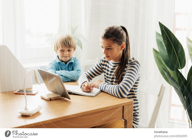 A schoolgirl, sitting at a table with her younger brother, performs an assignment on a tablet and makes notes in a notebook learning lesson together home video