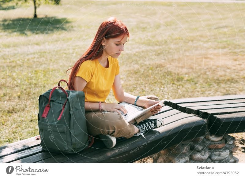 A teenage girl is sitting on a park bench with a laptop and preparing for lessons or exams. The concept of training and education backpack using student woman