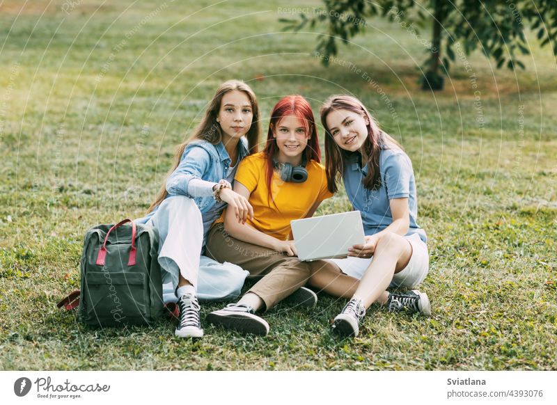 Teenage girls are sitting on a green lawn in a park with a backpack and a digital tablet and are preparing a joint project. Education and knowledge, lifestyle of teenagers