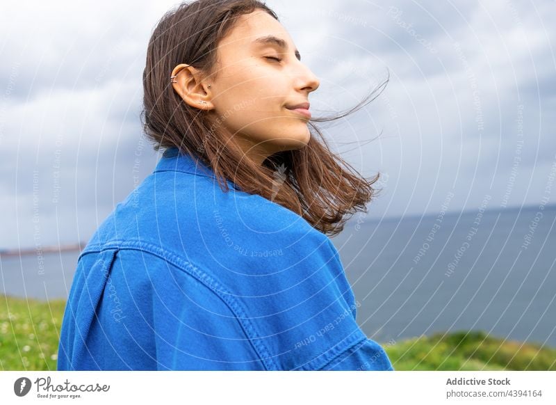 Content woman sitting on hill against sea eyes closed enjoy seaside cloudy admire smile seafront harmony female nature sky green ocean cheerful happy coast