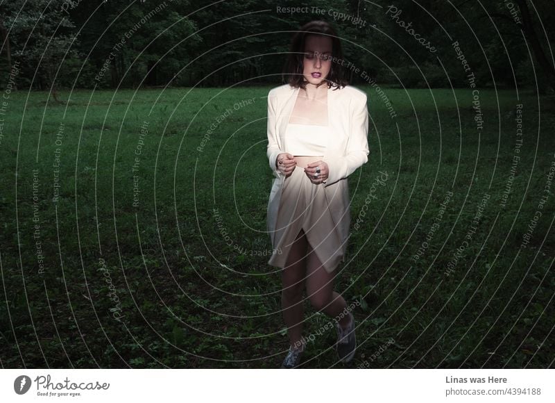 Green grass. White dress. A gorgeous brunette model is running all wild in these dark woods with a moody meadow. The avant-garde outfit is being enlightened during a photo session. It gives a mysterious feeling about all this scenery.