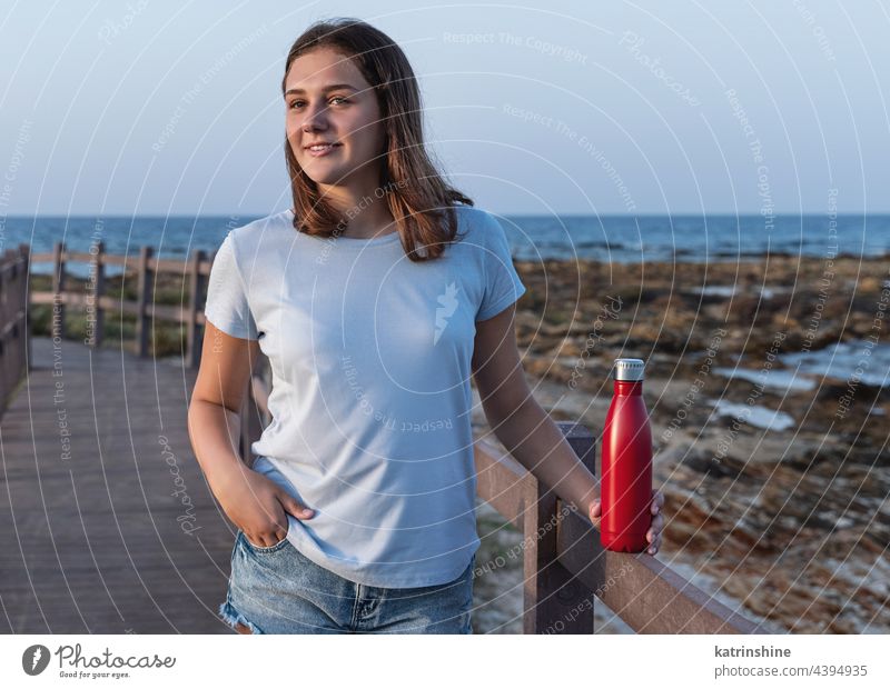 Teen girl in t-shirt standing and holding reusable water bottle in hands teen sea sunset mockup red blue light blue close up metal template jeans copy space