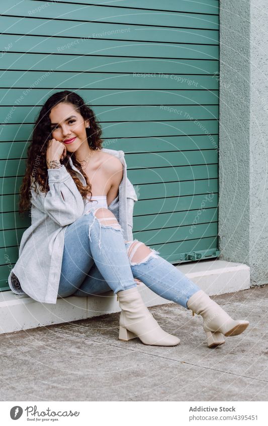 Glad woman sitting near building wall in city charming style smile street urban trendy fashion appearance female lean on hand cheerful glad outfit happy