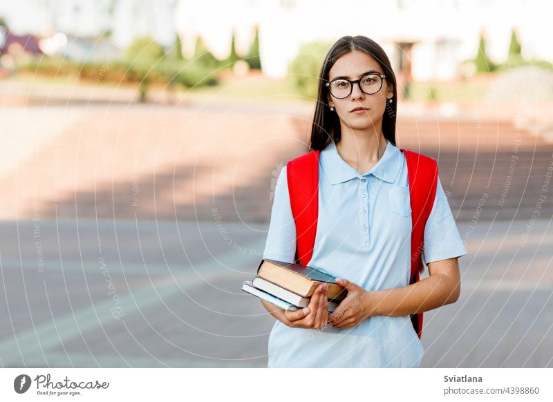 A smiling student girl with glasses, books and a red backpack. Portrait of a student on a city background. The concept of education college stair street