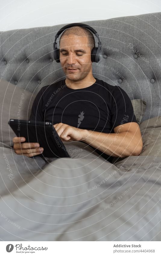 Man using tablet in bed in morning at home man browsing listen music bedroom awake male gadget rest device relax watch video surfing internet headphones