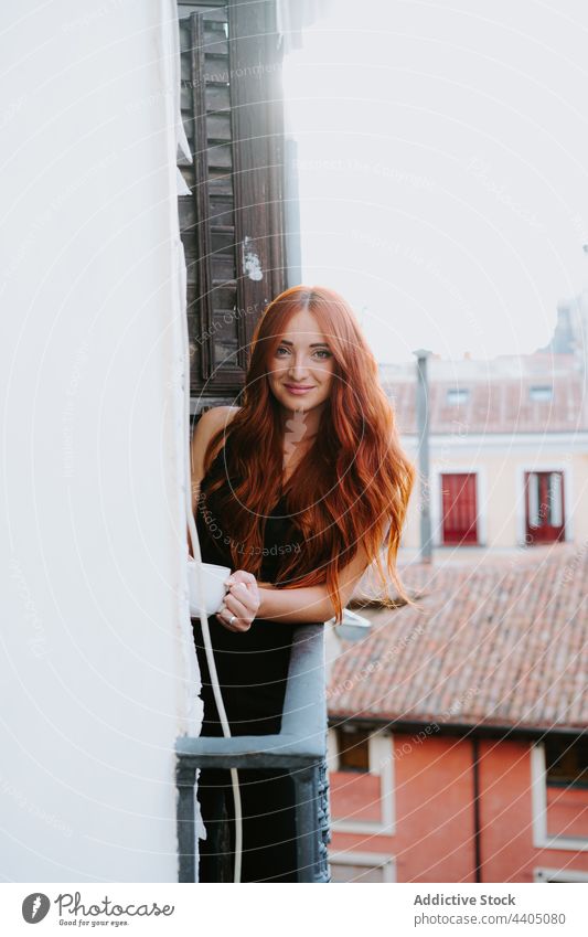 Smiling redhead woman with cup of drink on balcony beverage enjoy residential building smile female coffee tea chill content mug glad style fresh red hair