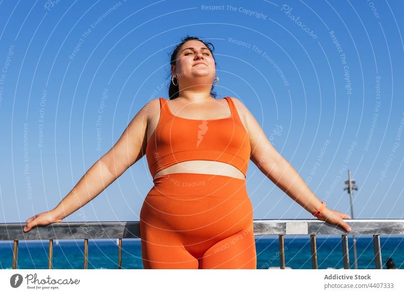 Smiling overweight ethnic sportswoman against fence and sea eyes closed smile dreamy enjoy idyllic plus size embankment blue sky ocean athlete break mindfulness