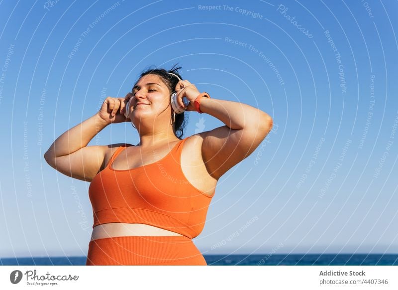 Smiling overweight ethnic sportswoman in headset under a blue sky athlete headphones listen music free time smile wall using gadget device cellphone wireless