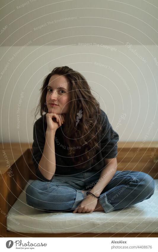 Portrait of a young woman sitting on a light-colored cushion, wearing jeans and a sweater, looking over at the camera while leaning on her hand Woman