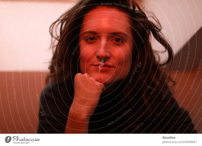 Portrait of a young woman sitting in red light, propping her chin with her fist and smiling at the camera Woman Young woman Brunette long hairs Long-haired