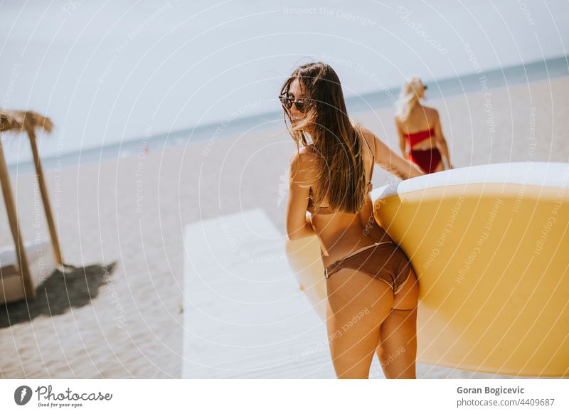 Two young women with paddle board on the beach on a summer day attractive beautiful bikini coast female fit friends friendship fun happy lifestyle ocean outdoor