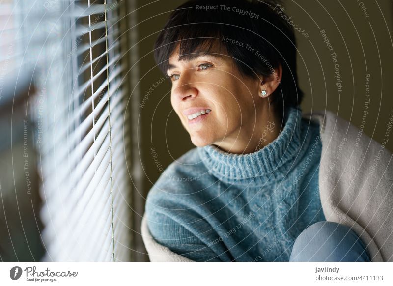 Happy middle-aged woman smiling while looking out the window. home 50s 40s female person brunette people interior smile relax white comfort indoor sweater