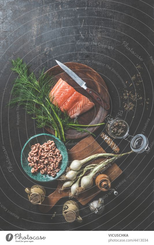 Raw salmon fillet on wooden plate with dill, knife and other ingredients for preparing a delicious fish dish: onions, shrimp on cutting board and jars of dry seasoning. View from above