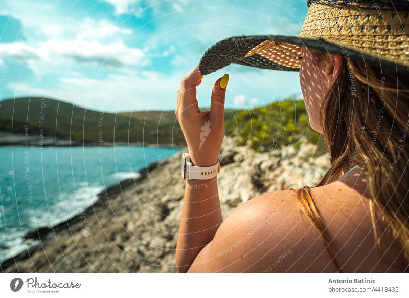 Side view of an unrecognizable brunette girl, holding onto her hay hat in the summer. Standing near the shore of the adriatic sea, concepts of travelling, summer and vacations