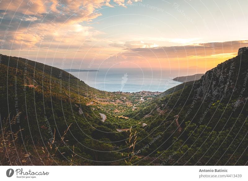 Amazing sunset on the Vis island in Croatia. Komiza city in the distance. Golden hour, orange sky and the beautiful blue adriatic sea. Green mountains surrounding the little picturesque town