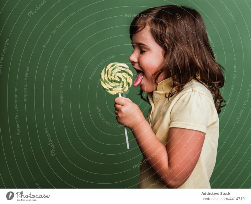 Cheerful girl with spiral candy on green background lick lollipop swirl sweet funny tongue out child kid dessert cute adorable happy tasty delicious stick treat