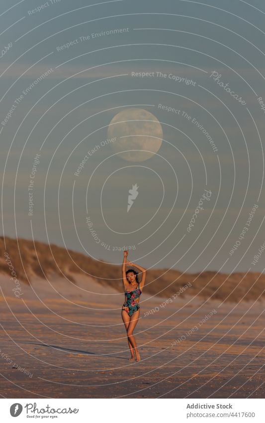 Graceful woman standing in seashore in sunset beach evening summer swimsuit serene twilight sand female moon sky swimwear idyllic arms raised sundown freedom