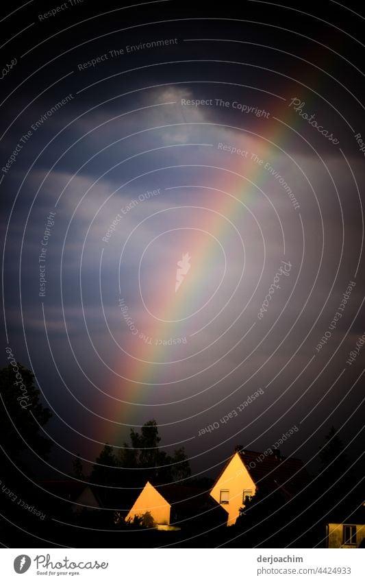 A rainbow meets from the dark cloud band, still on a somewhat blue sky. The two houses there shine in gold yellow colors from the setting sun, in front of the already dark trees.