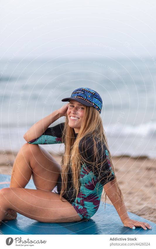 Cheerful woman sitting on SUP board on beach surfer paddleboard sup board swimsuit smile seashore summer female vacation swimwear hat recreation enjoy positive